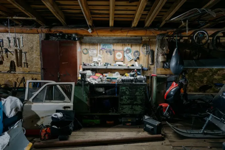 A cluttered garage filled with tools, motorcycle, and car door parts illustrating the consequences of delayed junk removal.