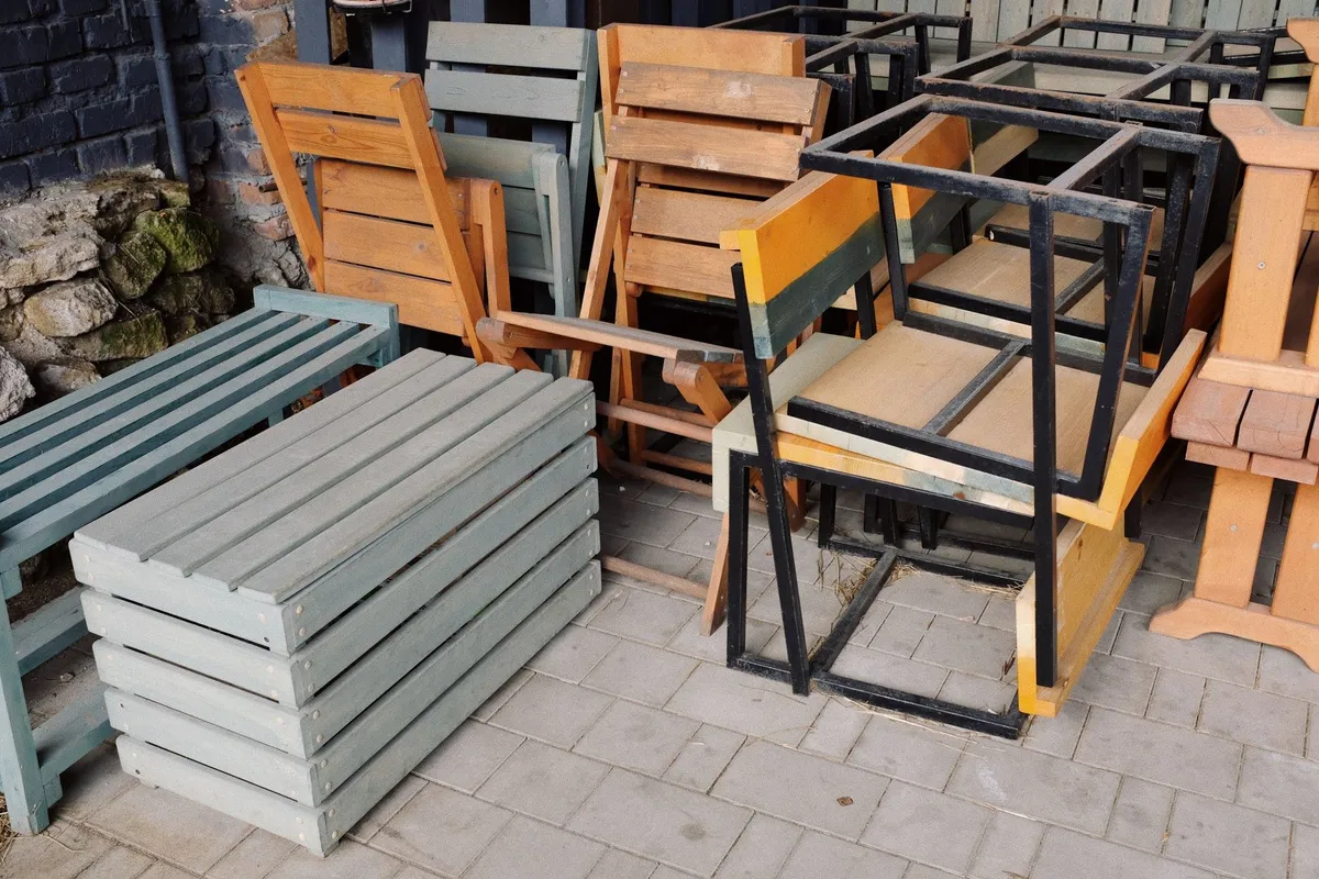 A variety of wooden benches and metal chairs stacked outside, suggesting it's time for furniture removal.